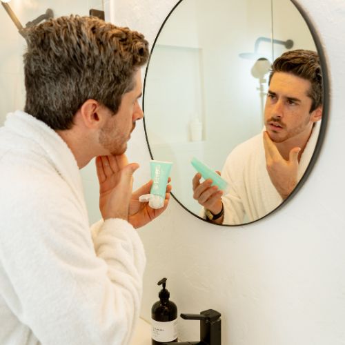 male model in a white bathrobe looking the mirror while applying revealu face moisturizer 